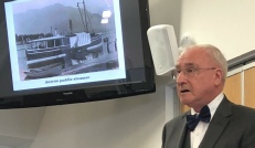 A man in a suit speaks beside a screen showing an old photograph of a lake steamer or launch