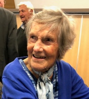 An elderly woman in a blue cardigan smiles at the camera among a small crowd