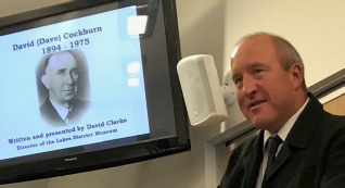 A man in a suit stands beside a screen showing a portrait of David (Dave) Cockburn, 1894-1975, written and presented by David Clarke
