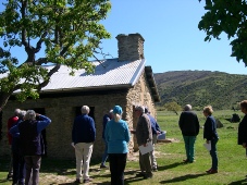Members gathered beside a restored stone cottage with a corrugated iron roof and a tall stone chimney, tussock hills behind