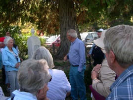 A guide addresses a group of visitors gathered outdoors near a stone cairn under trees