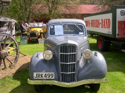A grey 1930s car with the registration JRK499 parked on grass at a vehicle gathering, a red truck and a horse-drawn wagon behind it