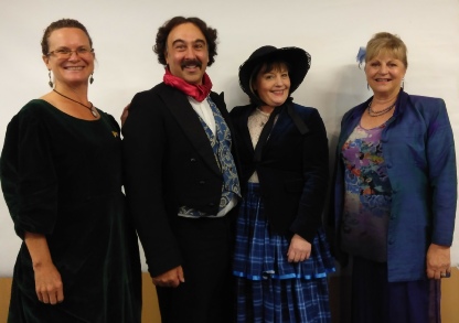 Four people standing side by side against a pale wall, two of them in period costume, a man in a black tailcoat with a red neckerchief and a woman in a feathered hat and tartan skirt, and two women in modern dress