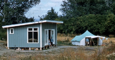 A small blue weatherboard crib on piles in rough grass, its door open, with a canvas tent pitched beside it and trees behind