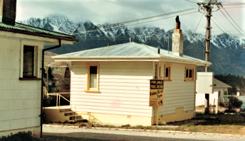 A cream weatherboard crib with a corrugated roof and a brick chimney beside a neighbouring house, the snow-covered Remarkables rising behind