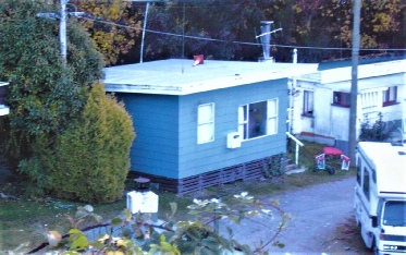 A blue weatherboard crib with a flat roof and a lean-to porch in a camp ground, a caravan and a truck parked alongside it in autumn