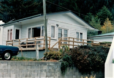 A cream weatherboard crib raised on a concrete block wall, with a timber deck and rail across its front and a dark car parked below
