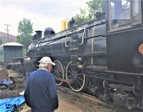 A man in a white cap kneeling at the side of a black steam locomotive under restoration, its driving wheels and rods in view