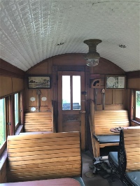 The interior of a wooden railway carriage, its varnished slat seats facing each other under a curved white ceiling and a brass lamp