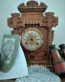 A carved wooden mantel clock with a painted dial, standing beside a leather-cased pair of binoculars, a handwritten document and a botanical print on a lace cloth
