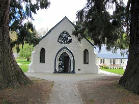 A small white weatherboard church with a steep gable and a pointed arch doorway, framed by two large conifers on a gravel path