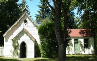 A white weatherboard church with a rose window above its pointed arch porch, a cream red-roofed hall beside it under a large summer tree