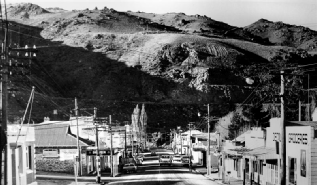 Black and white view down a main street of low shopfronts and power poles towards a church spire, with bare hills rising behind the town