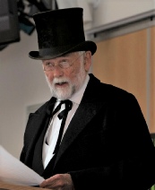 A bearded performer in a top hat, frock coat and white cravat reading from a sheet of paper