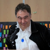 A performer in a dark jacket and a frilled white shirt with a blue brooch, speaking at a microphone