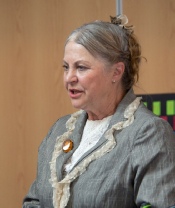 A performer in a grey period jacket with a fur-trimmed collar and her hair pinned up, speaking to the audience