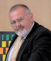 A bearded performer in glasses, a dark jacket and a gold waistcoat, looking towards the camera