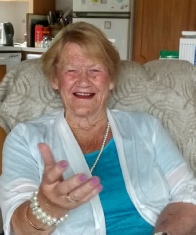 An older woman laughing in an armchair in a domestic kitchen, wearing a blue top, a white cardigan and a pearl necklace