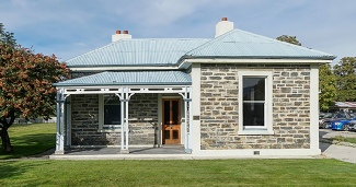 A restored single-storey stone cottage with a corrugated roof, two chimneys and a timber veranda, standing on a mown lawn