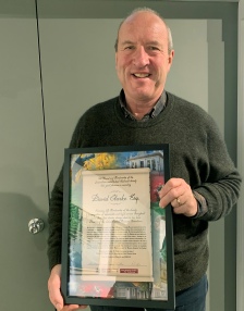 A smiling man in a dark green jumper holding up a framed illuminated Testimonial