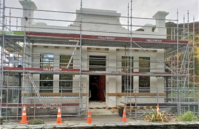 A two-storey classical stone building wrapped in Fallproof scaffolding, with road cones and a barrier across its doorway