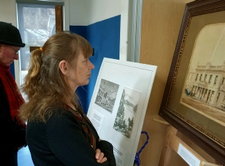 A woman in profile studying a mounted display of small framed drawings in a hall, with a large gilt-framed watercolour of a colonnaded building beside it