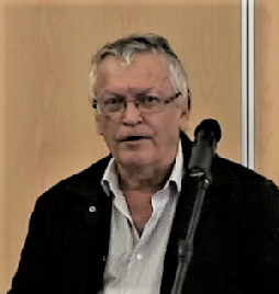 A man with grey hair and glasses in a dark jacket and open-necked shirt speaking at a microphone against a plywood wall