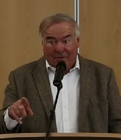 A man in a tweed jacket and open-necked shirt pointing towards the audience as he speaks at a lectern microphone