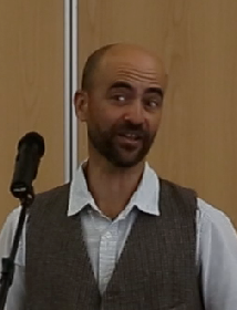A bearded man in a grey waistcoat and pale shirt speaking at a microphone, looking off to one side