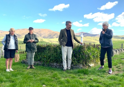 Four people standing at an iron-railed grave plot with a white cross, tussock hills and a blue sky behind, one of them speaking into a microphone