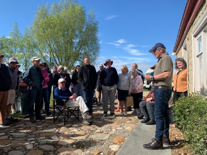 About twenty members gathered on a stone path beside a rendered mud-brick building while a man in a cap addresses them