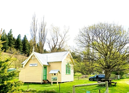 A small pale yellow weatherboard schoolhouse with green trim and a Cambrian School sign, an interpretation panel and a picnic table on the grass in front
