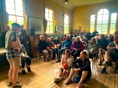 The interior of the Cambrian schoolroom, about thirty members seated on chairs and on the wooden floor under arched windows while a man in a cap teaches at the front