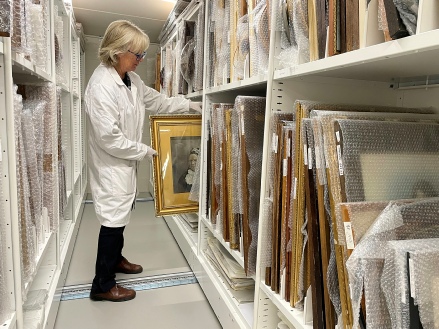 A woman in a white conservator's coat sliding a gilt-framed portrait out of racking in a museum collection store, bubble-wrapped pictures on the shelves either side