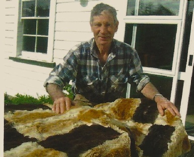 A man in a checked shirt standing against a white weatherboard wall behind a large rug of brown, white and black rabbit skins spread out in front of him