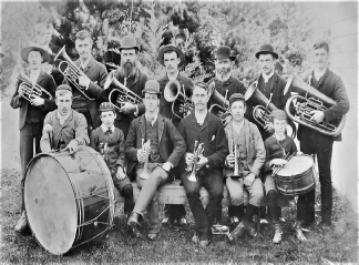 Black and white group portrait of about fifteen bandsmen and boys posed outdoors with cornets, tenor horns, a tuba and a large bass drum