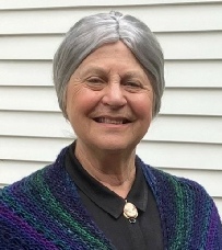 A smiling grey-haired woman in a green and blue striped cardigan and a brooch, standing outdoors against a weatherboard wall