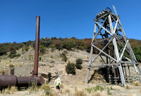 A timber mine poppet head on a dry hillside beside a rusted horizontal boiler and its tall chimney, a walker in the foreground