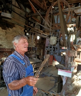 A man in a striped shirt and blue overalls holding a tool up to the light inside a belt-driven machine shop