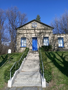 A small stone building with a pedimented gable, bright blue doors and blue window frames, reached by a concrete stairway with a handrail up a grassy bank