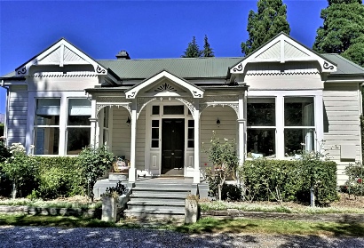 A cream weatherboard villa with a green roof, two bay windows and a fretwork porch, with garden beds and a lawn in front