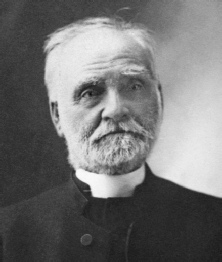 Black and white studio portrait of an elderly man with a white beard and a clerical collar, wearing a dark coat