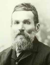 Black and white studio portrait of a man with swept back hair and a long beard, wearing a dark jacket and waistcoat
