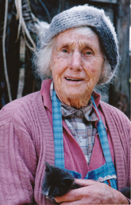 An elderly woman standing outdoors in a grey knitted hat and a pink cardigan over a blue checked apron, holding a small black kitten against her chest, with a rough shed wall behind her