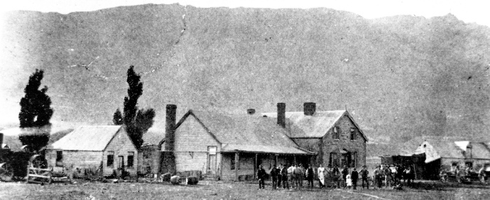 Black and white view of a row of single storey iron roofed buildings below a bare hillside, with about twenty people lined up outside them