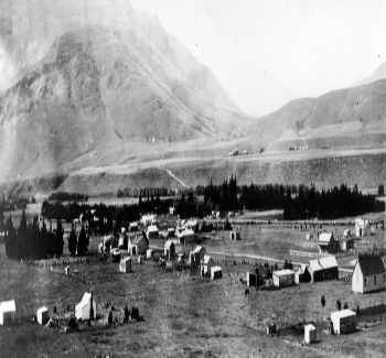 Black and white view across a valley floor scattered with small huts and tents, a steep mountain face rising behind