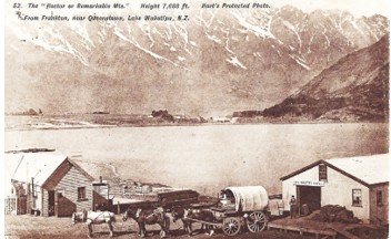 Sepia postcard view across a lake to bare mountains, with two small buildings, a covered wagon and horses on the shore in the foreground