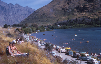 Colour photograph of a crowded lakeside beach with parked cars, sunbathers on a grassy bank and swimmers and small boats on the water below steep hills