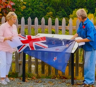 Two people at a picket fence lifting a New Zealand flag off a newly unveiled outdoor information panel, autumn trees behind them
