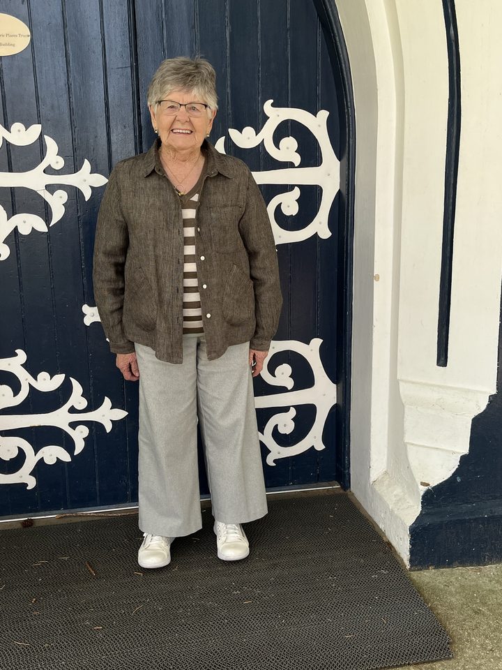 Mary May as herself, in a brown linen jacket and grey trousers, standing at a blue arched door with white ironwork